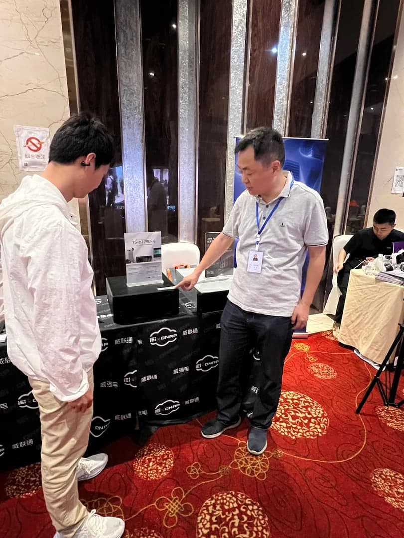 美步科技工程师在 2026 四川系统集成行业千人峰会威联通 QNAP 展位现场为系统集成商客户介绍全闪 NAS 企业存储产品,展位桌面摆放 QNAP NAS 设备实物,工程师手持产品宣讲